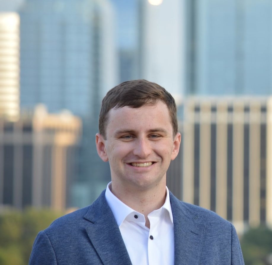 Ryan (student) smiles into the camera against a colorful blurred cityscape background.