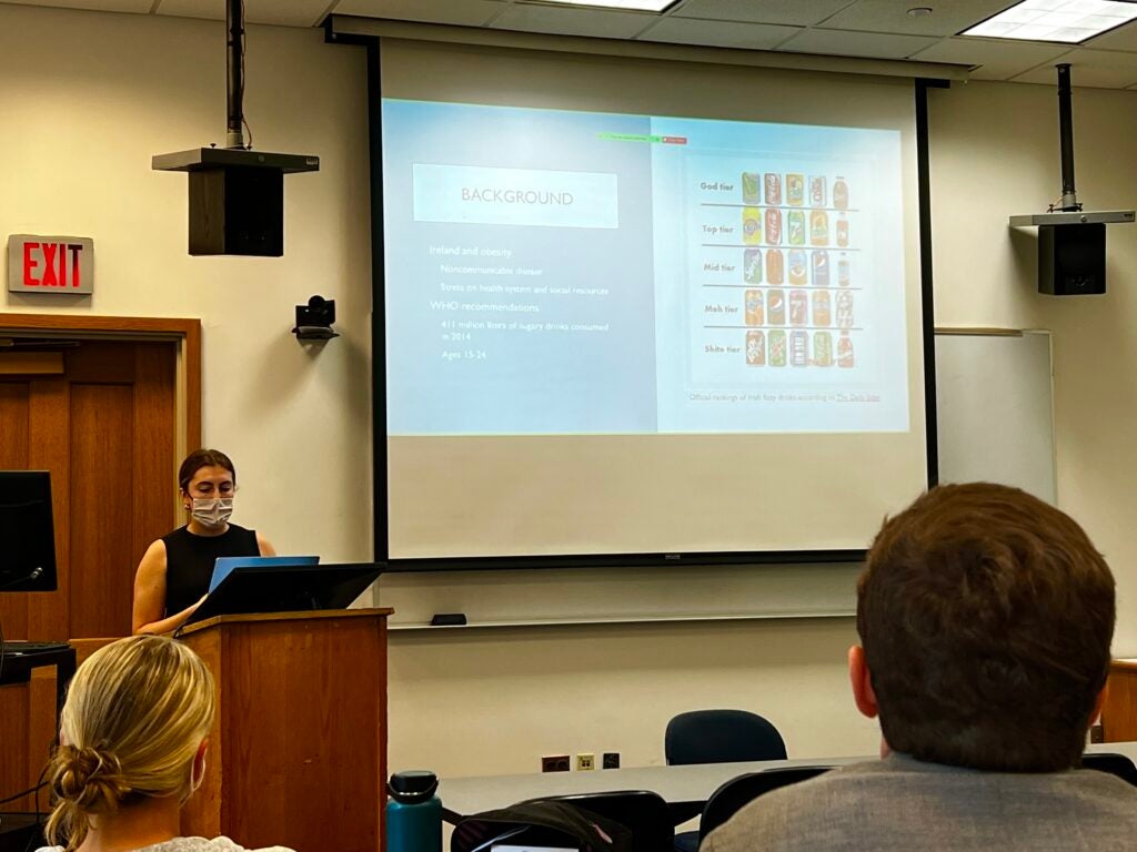 Siobhan Mitchell standing behind a podium. A powerpoint slide with soda bottles is displayed on the wall.