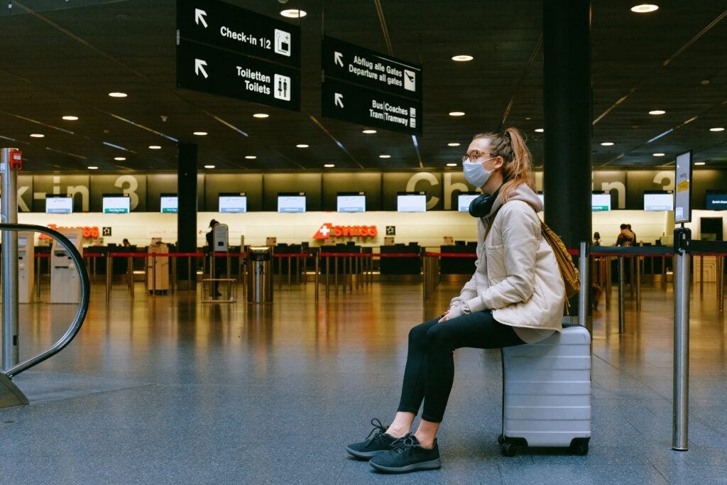 A traveler waits in the airport wearing a surgical mask, she sits on her suitcase and the room is otherwise empty.