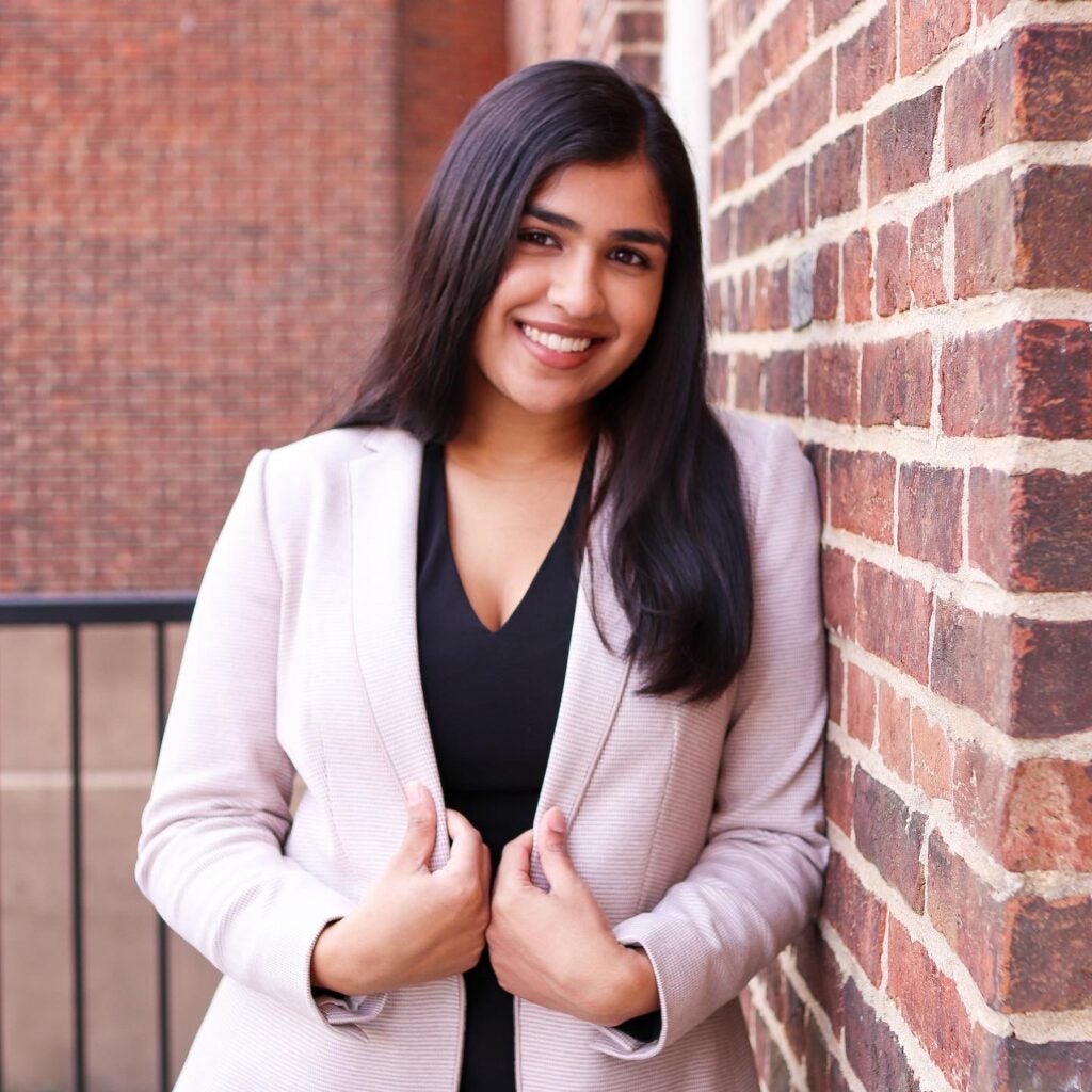 Alisha (student) smiles into the camera while leaning against a brick wall.