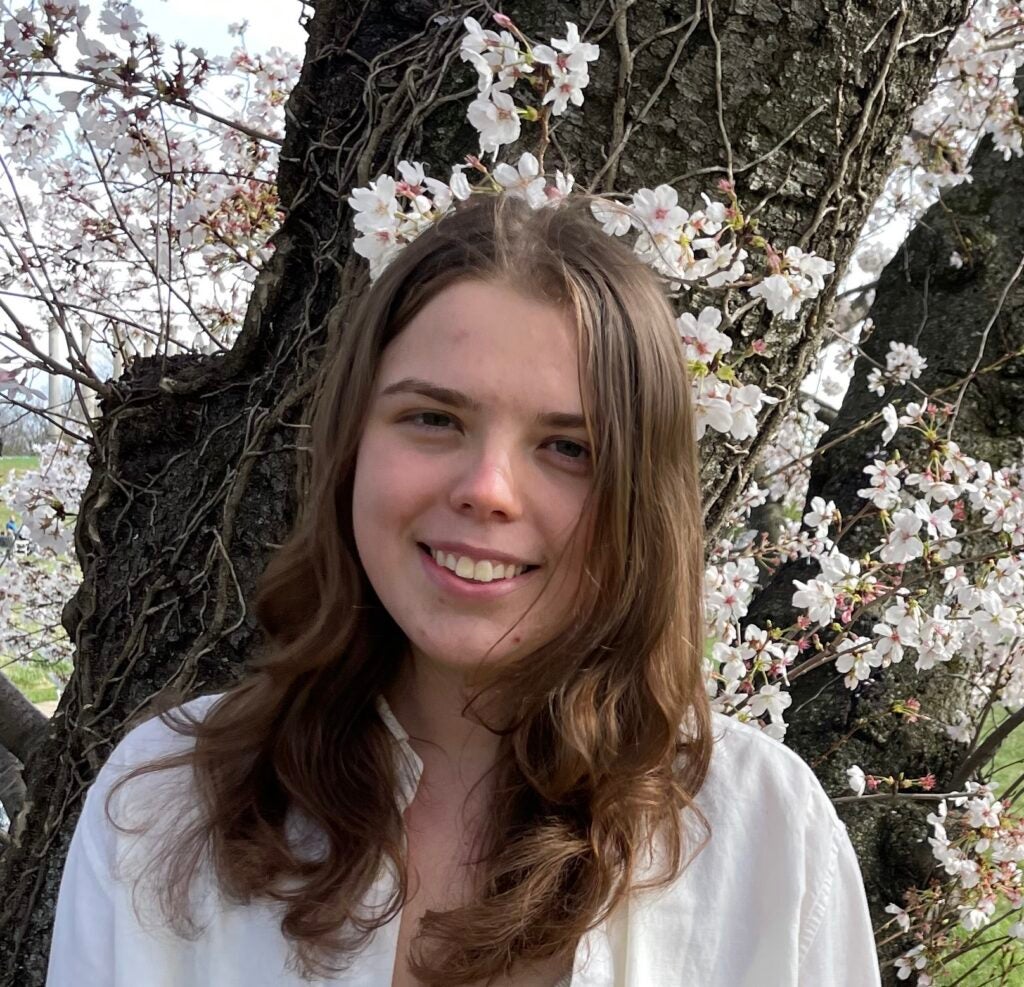 Maja (student) smiles into the camera while standing in front of a backdrop of pink cherry blossoms.