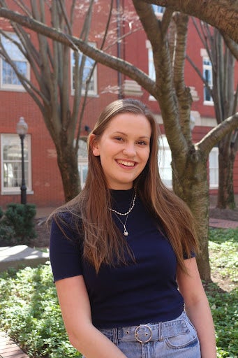 Madison Dwyer (student) smiles into the camera in front of a backdrop of trees and a red brick building.