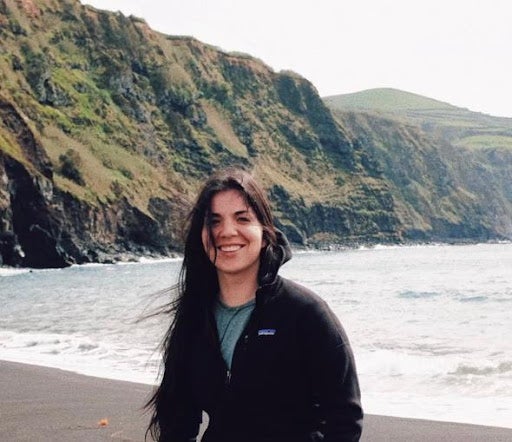 Sofia (student) smiles at the camera while standing in front of a mountain range and body of water.