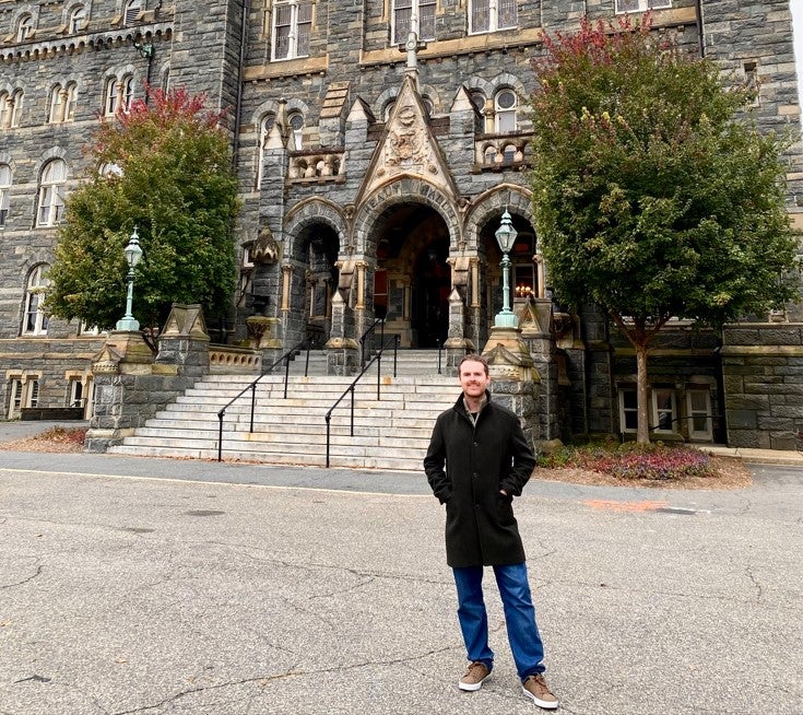 Dr. Darragh Gannon stands in front of Healy Hall. He wears a black trench coat and smiles at the camera.