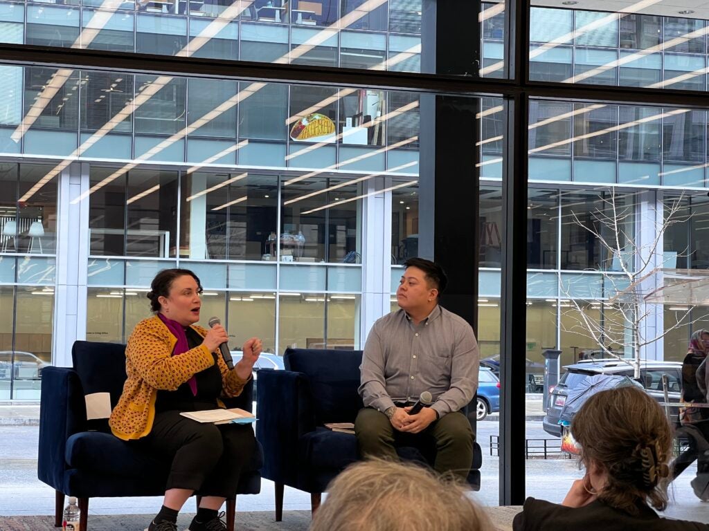 Jennifer Natalya Fink and Jayne A. Quan sit talking on stage against a glass wall overlooking the street