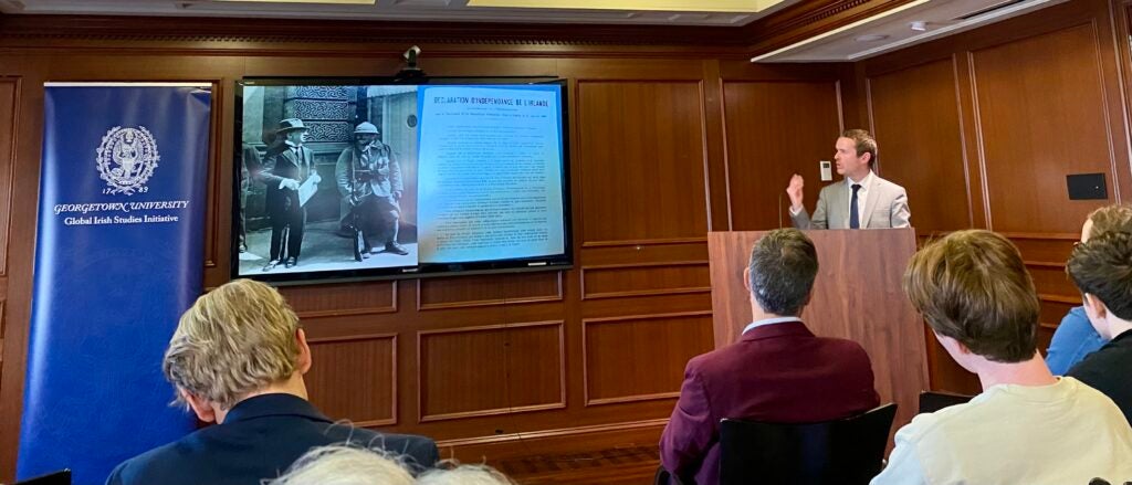 Dr. Darragh Gannon speaks at a podium in front of a screen showing a black and white photo.