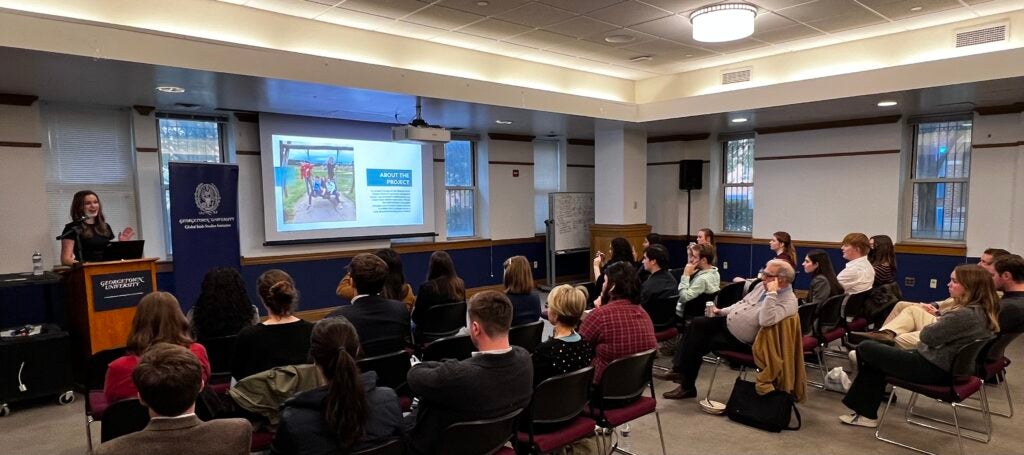 a crowd of people sit in rows watching a student at a podium speak.