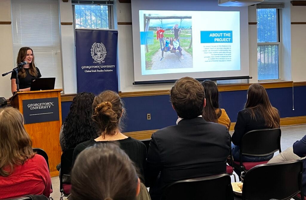 a crowd of people sit in rows watching a student at a podium speak.