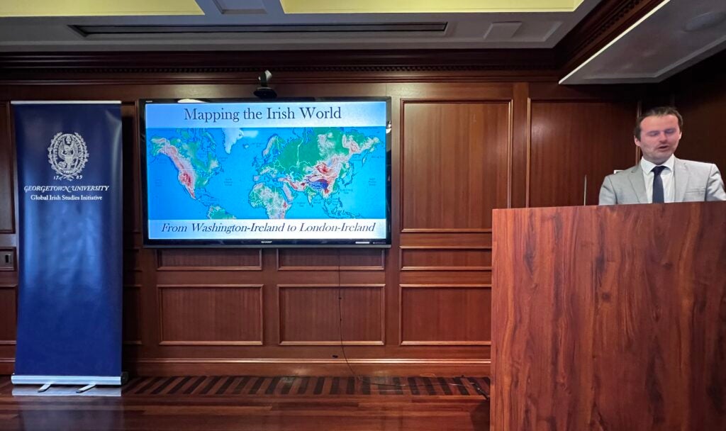 Dr. Darragh Gannon speaks at a podium in front of a screen reading "Mapping the Irish World"