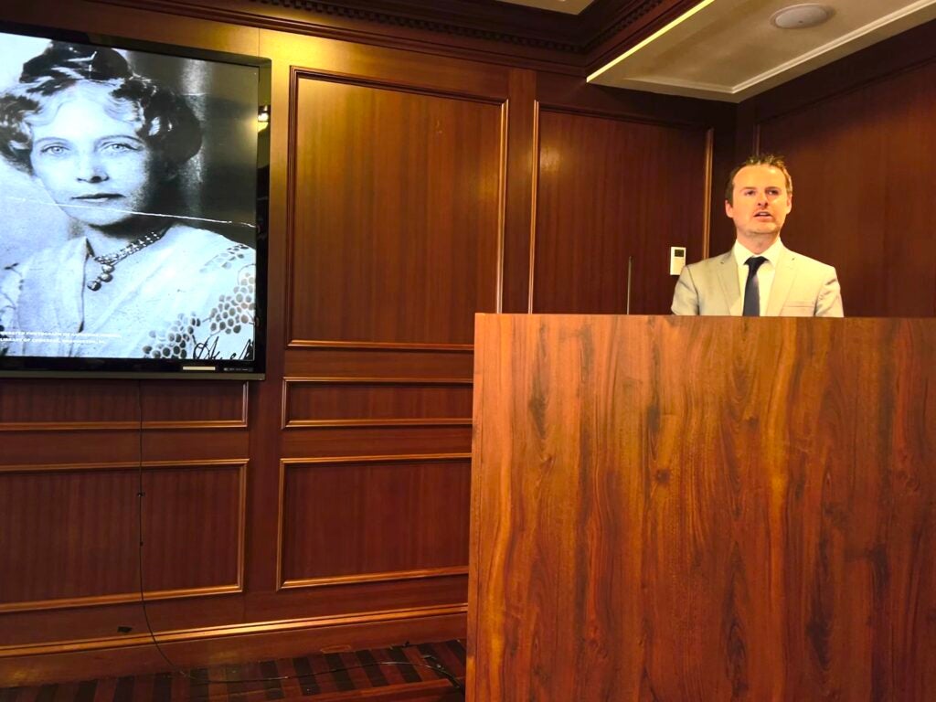 Dr. Darragh Gannon speaks at a podium in front of a screen showing a black and white photo of Katherine Hughes.