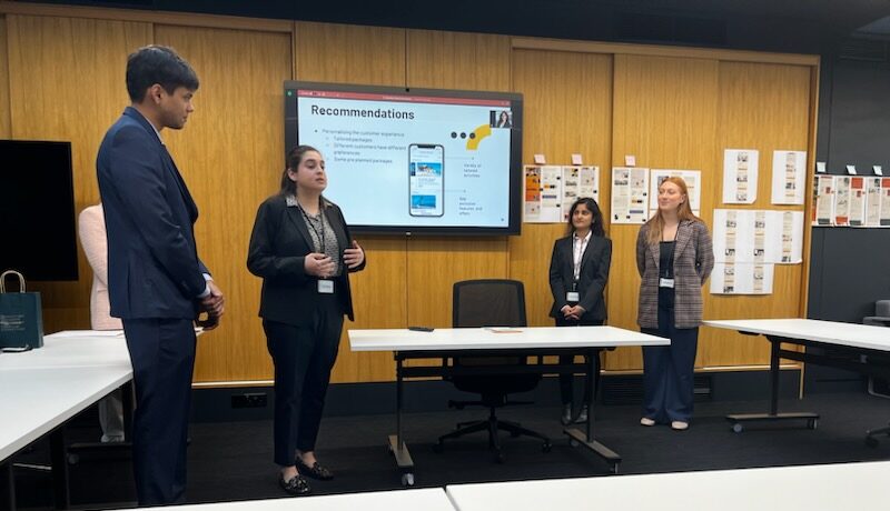 four students stand in the front of a meeting room with a presentation behind them reading "recommendations"
