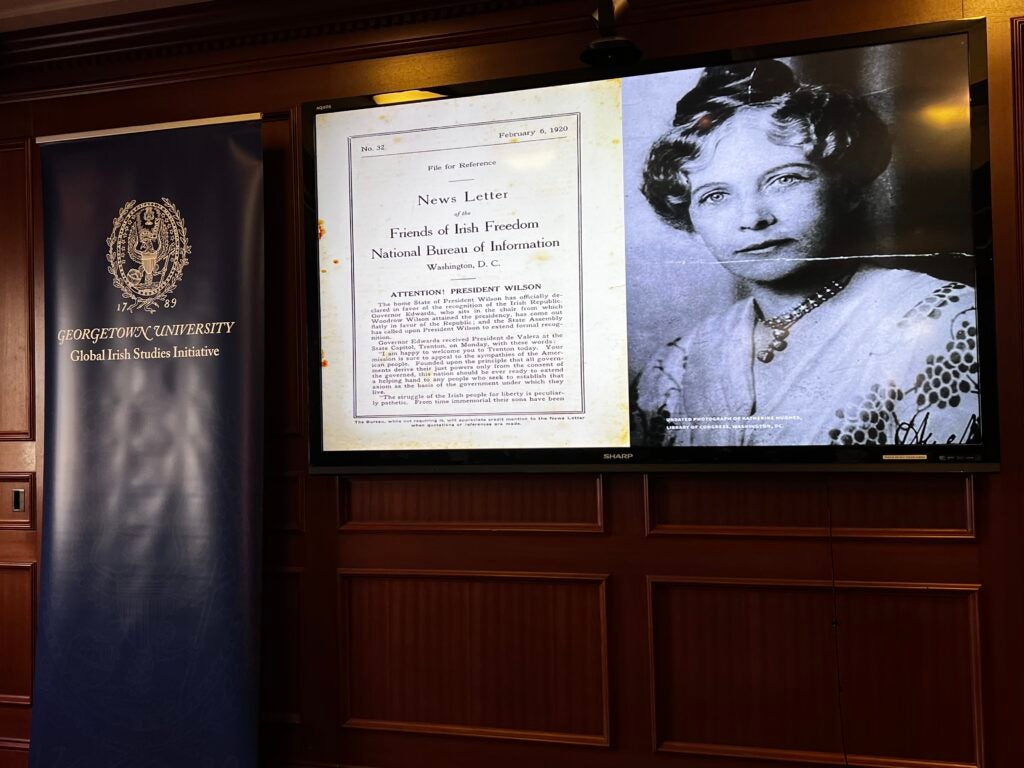 A screen shows a newsletter from 1920 beside a black and white photo of Katherine Hughes. A navy Global Irish Studies Initiative banner is beside the screen.