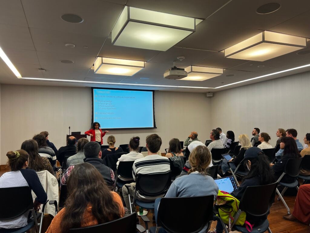 image is of a large seminar room with white walls. the front of the room has a drop down screen presenting the speaker's powerpoint. There is a large audience in front of screen of about 50 people. Laia Balcells stands in the front in an orange sweater and black trousers.