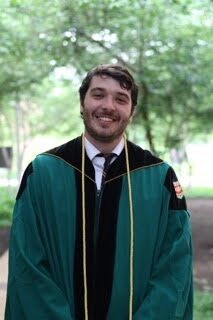 Picture of a smiling person wearing a green graduation robe with a yellow cord