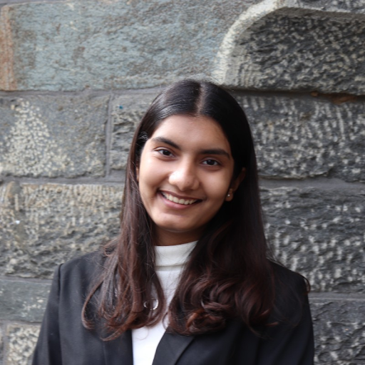 Picture of a smiling person in a black blazer and white turtleneck with a stone background